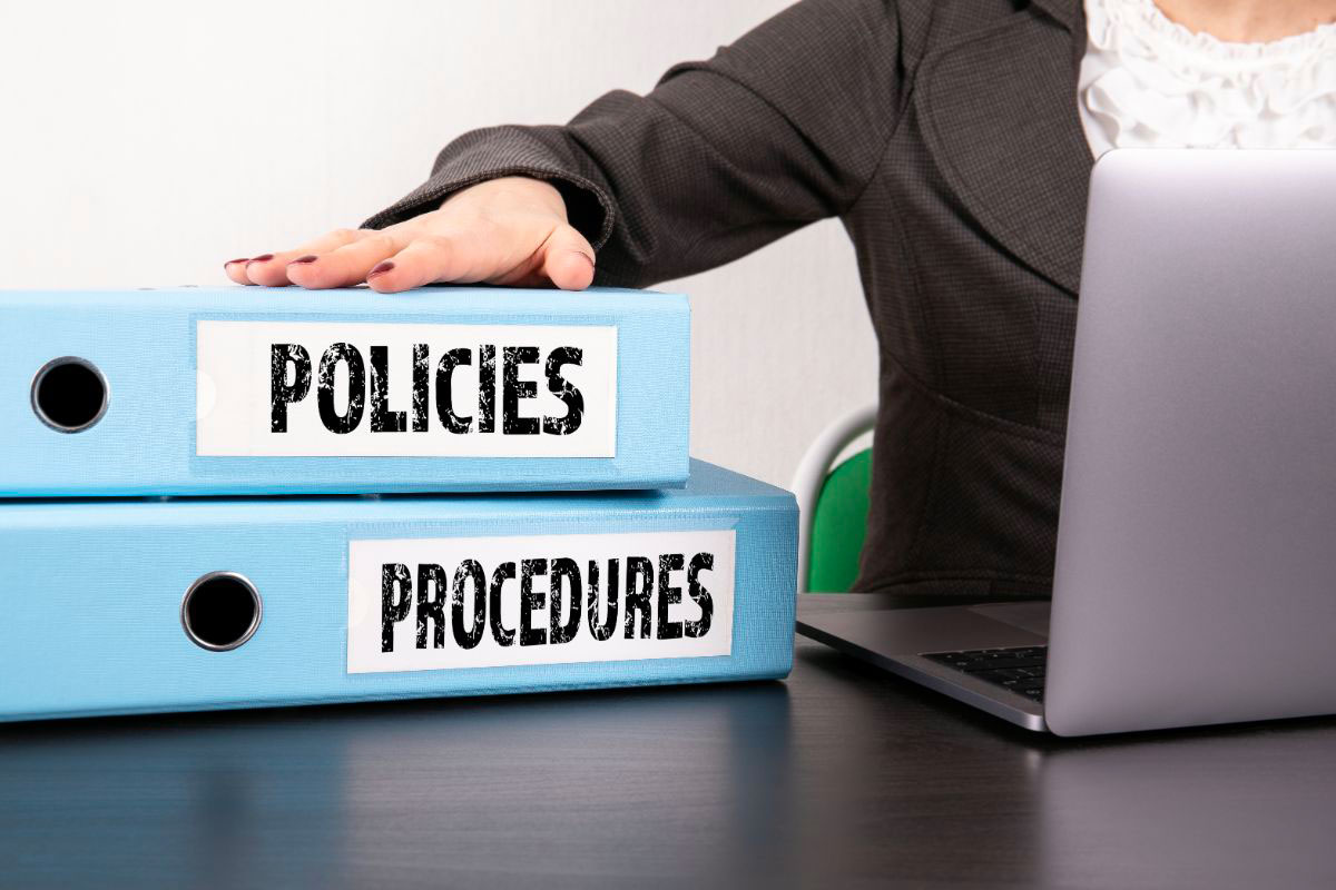 A hand resting on two binders labelled ‘Policies’ and ‘Procedures’, symbolising the management and organisation of compliance and regulatory policies and procedures.